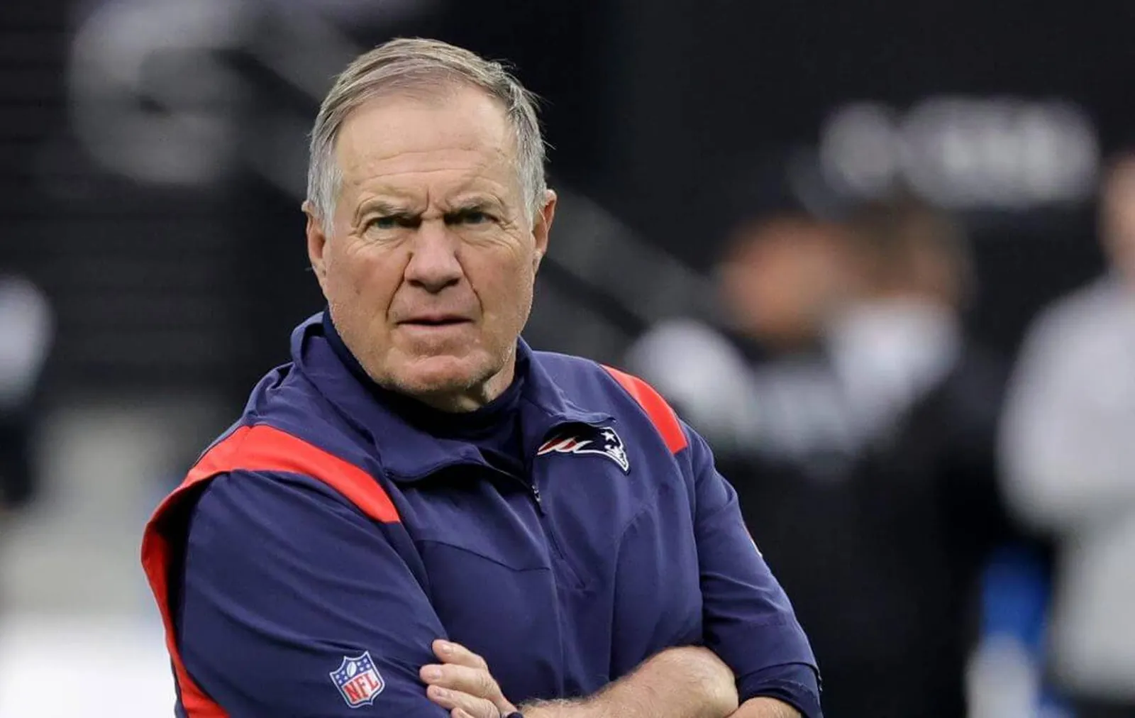 Bill Belichick, wearing a navy Patriots jacket, folds his arms and looks forward with his brow furrowed.